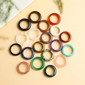 May include: A collection of twenty circular rings crafted from various materials, including solid black, white, red, green, blue, and translucent stones, as well as speckled, glittery, and marbled patterns arranged in a scattered formation.