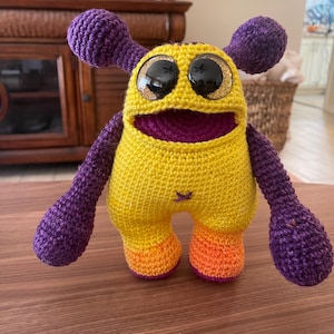 Puede incluir: Un juguete de monstruo de ganchillo hecho a mano en amarillo, morado y naranja. El juguete tiene ojos grandes y brillantes, una boca ancha y dos antenas moradas. Los brazos y las piernas también son morados y los pies son naranjas.