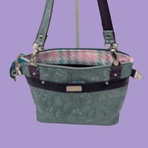 May include: A teal handbag featuring a subtle floral pattern, a dark purple horizontal band, and matching dark purple shoulder straps. The interior is lined with a light-colored plaid fabric. The bag includes silver-toned hardware and a zipper closure.