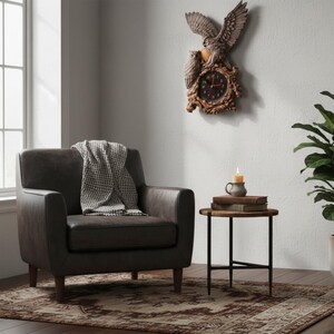 May include: A wall clock featuring a sculpted owl with spread wings perched above a smaller owl, surrounding a dark circular clock face with red hands. The clock is mounted on a wall above a dark grey armchair with a patterned throw blanket.