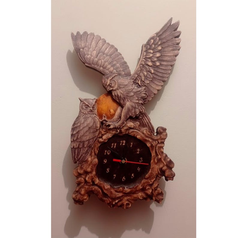 May include: A wall clock featuring two sculpted owls in shades of brown and grey. One owl is perched while the other has wings spread wide above a circular clock face with black background, gold numerals, and red hands, set in a textured frame.