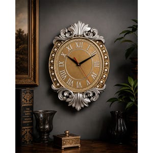 May include: An oval wall clock featuring a gold-toned face with black Roman numerals and thin black hands. The frame is finished in a metallic silver with ornate, scrolled detailing at the top and bottom, and a Greek key pattern border.