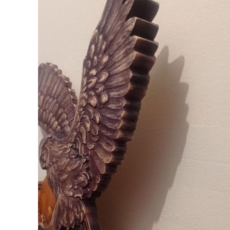 May include: A carved wooden wall sculpture depicting an eagle with wings spread wide. The wood features a dark brown finish with lighter highlights on the textured feathers, emphasising the depth and intricate detail of the avian form.