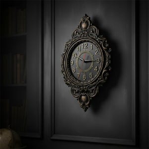 Può includere: Un orologio da parete decorato con una finitura metallica scura e dettagli a volute complessi. Il quadrante presenta uno sfondo nero con numeri arabi dorati, un motivo centrale a raggiera e tre lancette sottili, inclusa una lancetta dei secondi rossa.