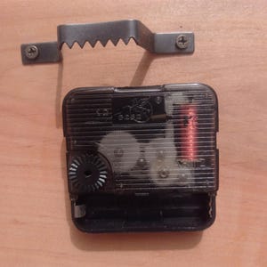 May include: A black plastic quartz clock movement mechanism with visible internal gears and a copper coil, accompanied by a separate metal sawtooth wall hanger bracket featuring two small screws for mounting purposes.