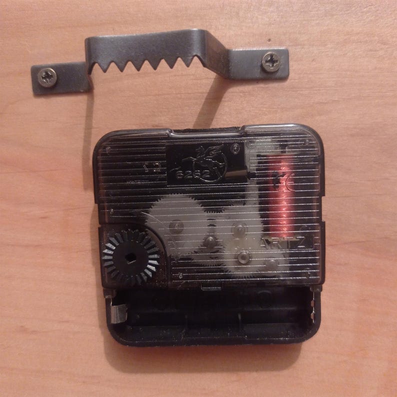 May include: A black plastic quartz clock movement mechanism with visible internal gears and a copper coil, accompanied by a separate metal sawtooth wall hanger bracket featuring two small screws for mounting purposes.