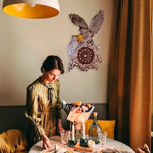 May include: A wall clock featuring a sculpted grey owl with outstretched wings perched above a second owl, both positioned over a dark brown circular clock face with white numbers, all set against a textured, wood-like decorative frame.