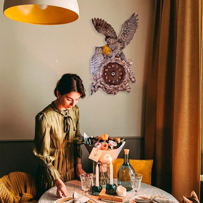 May include: A wall clock featuring a sculpted grey owl with outstretched wings perched above a second owl, both positioned over a dark brown circular clock face with white numbers, all set against a textured, wood-like decorative frame.