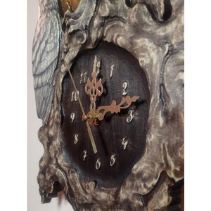 May include: A wall clock featuring a dark brown circular face with white Arabic numerals and ornate gold-coloured hands. The clock is encased in a textured, weathered grey frame with intricate relief carvings, including a prominent owl figure.