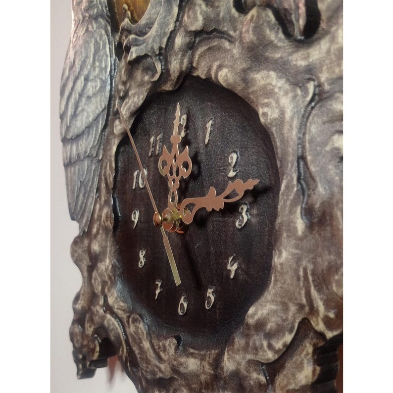 May include: A wall clock featuring a dark brown circular face with white Arabic numerals and ornate gold-coloured hands. The clock is encased in a textured, weathered grey frame with intricate relief carvings, including a prominent owl figure.