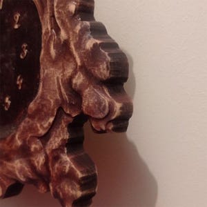 May include: A wall clock with a dark brown, textured wooden frame featuring a carved, irregular edge. The clock face is dark brown with visible light-coloured numbers. The wood grain shows a distressed finish with lighter highlights on the edges.
