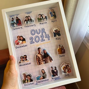 Peut inclure: Un présentoir photo encadré blanc avec le texte "OUR 2024" et des photos représentant chaque mois de l'année. Les photos sont de couples. Le présentoir est tenu par une main.