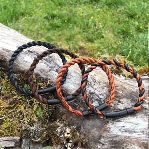 May include: Three braided leather bracelets with black and brown leather. The bracelets are on a wooden log with moss and green plants.