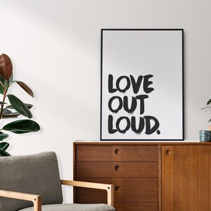 Könnte beinhalten: Ein gerahmter Kunstdruck mit den Worten "LOVE OUT LOUD" in fetten, schwarzen Pinselstrichen. Der Druck befindet sich auf einem Holzschrank, neben einer Topfpflanze und einem grauen Sessel. Links ist eine große Blattpflanze.