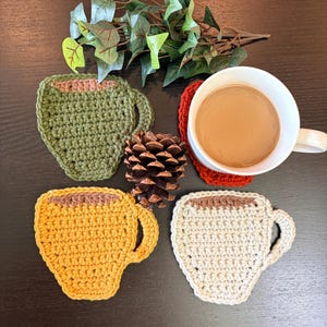 May include: A white mug filled with coffee sits on a red crocheted coaster. Three crocheted cup-shaped coasters in green, yellow, and cream are also visible. A pinecone and greenery complete the arrangement. The coasters are handmade.