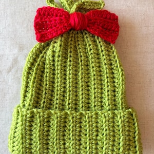 May include: A lime green crocheted beanie hat featuring a ribbed texture and a prominent, bright red crocheted bow attached to the top. The hat has a folded brim at the base and a small, knotted green crochet detail extending from the center top.