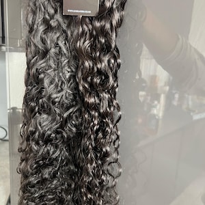 Pode incluir: Extensões de cabelo encaracolado castanho escuro. O cabelo é muito encaracolado e parece ser de cor natural. Uma etiqueta com as letras "LL" e o endereço do site www.luxlayers.co.uk está presa ao cabelo.