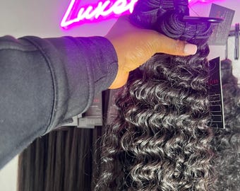 Pode incluir: Um feixe de extensões de cabelo encaracolado escuro. O cabelo é segurado por uma mão, com um letreiro de néon no fundo que diz "Luxel". As extensões de cabelo têm uma textura natural e ondulada e são de uma cor preta profunda.