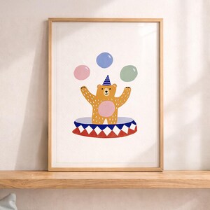 May include: A framed print featuring a whimsical illustration of a bear juggling three colorful balloons. The bear, wearing a party hat, stands on a circus ring with a red, white, and blue pattern. The artwork is displayed on a wooden shelf.