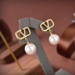 Op de afbeelding: Een paar goudkleurige oorbellen met een V-vormig ontwerp en een bungelende witte parel. De oorbellen worden gepresenteerd op een grijze standaard tegen een vage bruine achtergrond. Een klassiek sieraad.