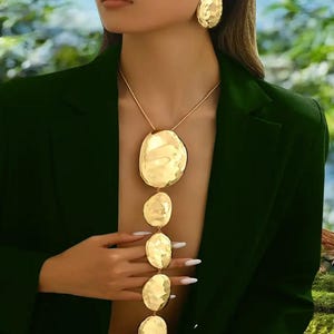 May include: Gold-toned jewelry set featuring a necklace with five textured, oval pendants and matching earrings. The necklace hangs from a delicate chain. The earrings are large, round, and textured. The jewelry is worn with a dark green blazer.