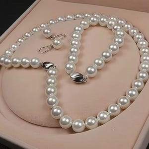 May include: A white pearl necklace and matching earrings set. The necklace features large, round pearls strung together with a silver clasp. The earrings have a single pearl on a hook. The set is displayed in a pink jewelry box.