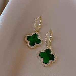 May include: A pair of gold-colored hoop earrings with a four-leaf clover design. Each clover is a deep green color, surrounded by a border of small, clear crystals. The earrings are displayed on a neutral-toned background.