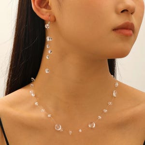 May include: A clear, beaded necklace and matching earrings set. The necklace and earrings feature clear, irregularly shaped beads strung on a nearly invisible thread. The earrings dangle from the ear, while the necklace rests on the collarbone.