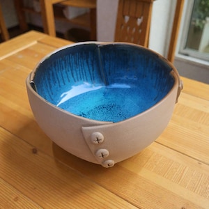 May include: A handcrafted ceramic bowl with a pale beige exterior and a striking blue interior. The bowl has a distinctive design, with three button-like embellishments on the side. The interior has a glossy finish with a dripped paint effect.