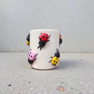 Puede incluir: Una taza de cerámica color crema decorada con figuras de mariquitas de colores. Las mariquitas son rojas, amarillas, rosas y negras con lunares y antenas detalladas. La taza es cilíndrica y se apoya sobre una superficie gris claro.