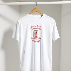 Op de afbeelding: Wit T-shirt met de tekst "Just a little something to take the edge off" in rode letters. Een illustratie van een Diet Coke-blikje staat in het midden van het shirt. Het T-shirt hangt aan een houten hanger.