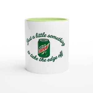 Puede incluir: Taza de cerámica blanca con borde interior verde. La taza presenta una imagen verde y blanca de una lata de Mountain Dew, con el texto "Just a little something to take the edge off" en escritura verde.
