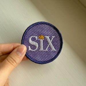 Puede incluir: Un parche redondo bordado con la palabra "SIX" en blanco. El parche tiene un fondo morado y un borde azul oscuro. Una pequeña corona dorada se encuentra encima de la "X".
