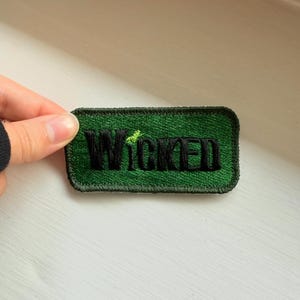 May include: A rectangular green embroidered patch with the word "WICKED" in black block letters. The patch has a black border and a small green accent above the "I".