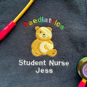 Könnte beinhalten: Marineblaues Fleece mit gesticktem Text und einem Teddybären-Design. Der Text lautet "Paediatrics" in Regenbogenfarben und "Student Nurse Jess" in Weiß. Ein Stethoskop mit rosa Griff ist im Bild.