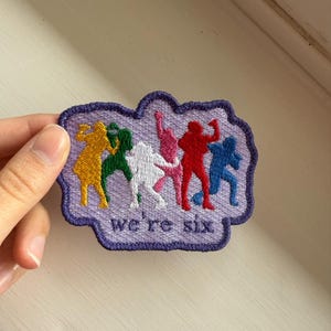 May include: Embroidered patch with a purple border and the text "we're six." The patch features six stylized figures in yellow, green, white, pink, red, and blue. The figures are in various dynamic poses.