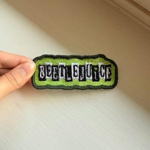 Puede incluir: Parche bordado con la palabra "BEETLEJUICE" en letras negras y blancas. El parche tiene un fondo verde brillante y un borde negro. El parche es rectangular con bordes redondeados.