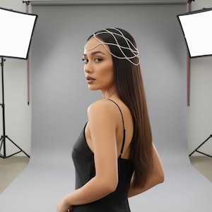 May include: A woman wearing a silver headpiece with a delicate, interwoven design of small, sparkling stones. The headpiece sits atop her head, with strands cascading down. She wears a black dress with thin straps. The background is a studio setting with soft lighting.
