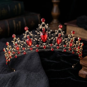 Op de afbeelding: Een goudkleurige kroon met rode en zwarte edelstenen. De kroon heeft grote, druppelvormige rode edelstenen en kleinere zwarte accenten. Het ontwerp is sierlijk en koninklijk, geschikt voor speciale gelegenheden of kostuums.