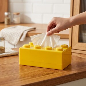 May include: A bright yellow tissue box designed to resemble a large building block. The box has yellow circular studs on top and is sitting on a wooden surface. A hand is pulling a white tissue from the box.