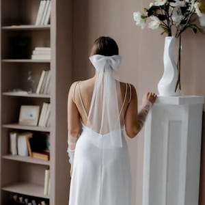 Könnte beinhalten: Eine Braut in einem weißen Hochzeitskleid mit tiefem Rücken und Spaghettiträgern. Sie trägt eine weiße Schleife und einen Schleier im Haar. Sie steht in der Nähe einer weißen Vase mit weißen Blumen.