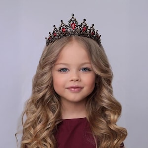 May include: A young person with long, wavy blonde hair and blue eyes wearing a dark crown with red gemstones. The crown is ornate and sits atop the person's head. The person is wearing a dark red top.