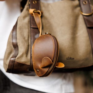 Può includere: Un portamonete a forma di balena in pelle marrone, con chiusura a zip e un piccolo anello in pelle. Il portamonete è appeso a un cinturino in pelle marrone attaccato a una borsa di tela con dettagli in pelle marrone.