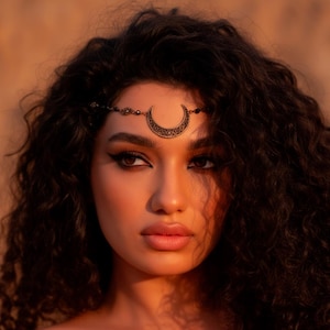 Pode incluir: Uma mulher com cabelo escuro e encaracolado usando uma tiara dourada em forma de lua crescente. A tiara é adornada com pequenos elementos decorativos. A pele da mulher tem um tom quente e dourado, e ela tem maquiagem escura nos olhos.