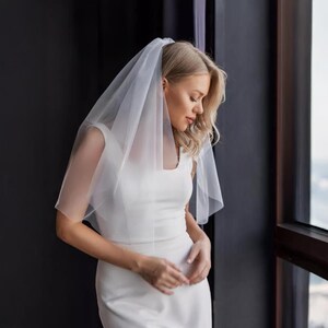 以下が含まれることがあります： 白いウェディングドレスと透け感のある白いベールを身に着けた花嫁。 ベールは頭と肩にかけられています。 花嫁は、暗いフレームの窓の近くに立っています。 ドレスはノースリーブで、スクエアネックです。
