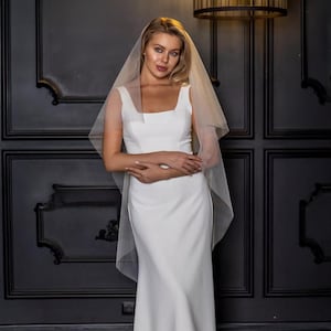 May include: A bride wearing a white wedding dress with a square neckline and a sheer veil. The dress is floor-length and form-fitting. The veil is draped over the head and shoulders. The background is a dark wall with decorative panels.