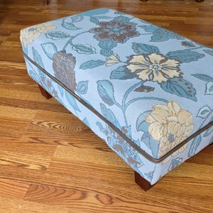 May include: A rectangular ottoman with a floral pattern in shades of blue, brown, and yellow. The fabric is trimmed with a brown border. The ottoman has four short, dark brown wooden legs.
