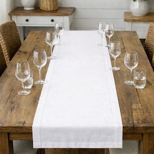 May include: A long, rectangular white table runner made of fabric with a hemmed border, centered on a rustic wooden dining table. The table is set with several clear glass wine glasses and small water tumblers arranged along the sides.