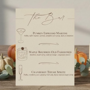 May include: A bar menu with three cocktail recipes: Pumpkin Espresso Martini, Maple Bourbon Old Fashioned, and Cranberry Thyme Spritz. The menu is on a wooden table with pumpkins, candles, and greenery.