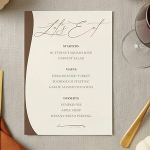May include: A fall-themed tablescape featuring a menu card with the text "Let's Eat", surrounded by white pumpkins, a gold spoon, a wine glass with red wine, and gold cutlery on an orange napkin.
